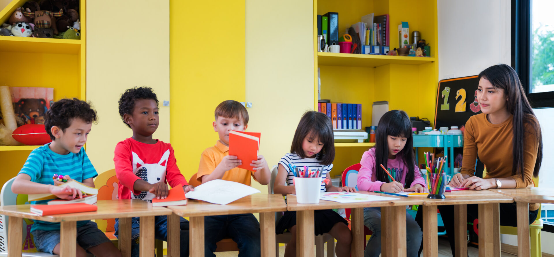 a teacher sits with a class of children at a desk as they read from books and draw