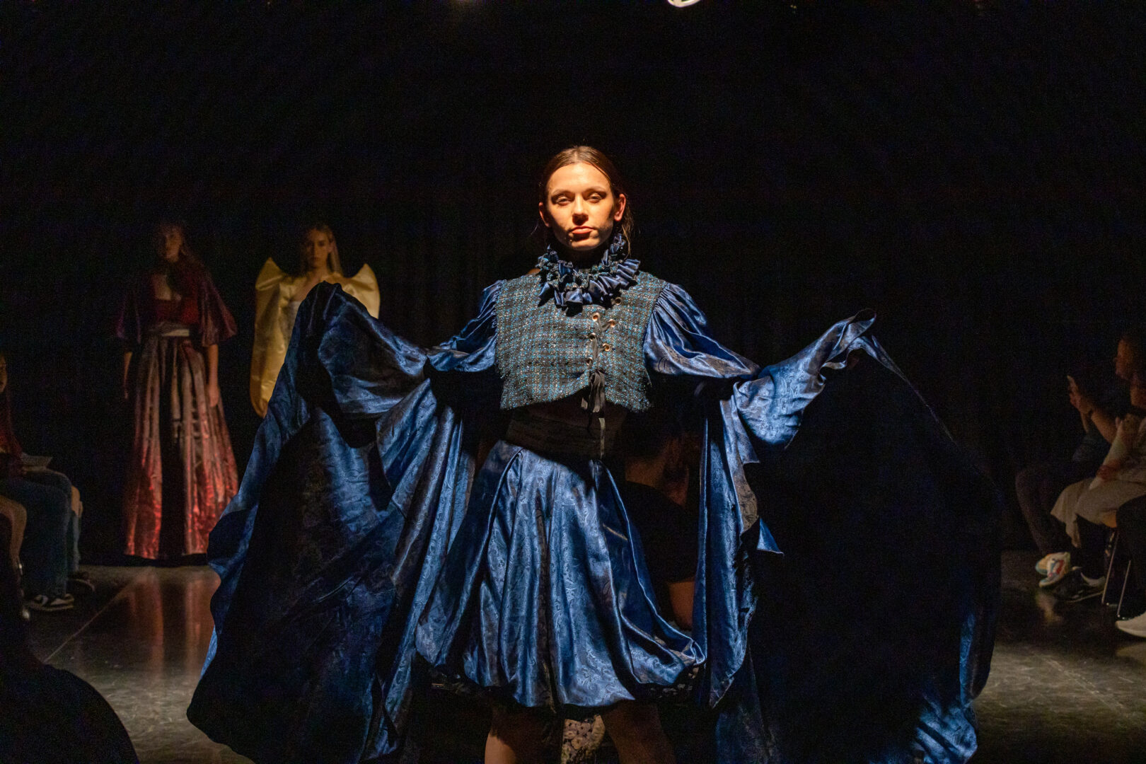 a model stands looking at the camera wearing a blue satin jacket and skirt as they hold the draped sleeves out by their side